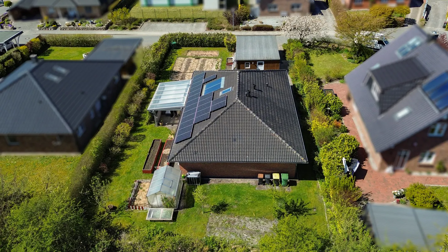 Aerial view of a brick house with solar panels on a dark gray roof, a greenhouse, and green bins in a lush green yard.