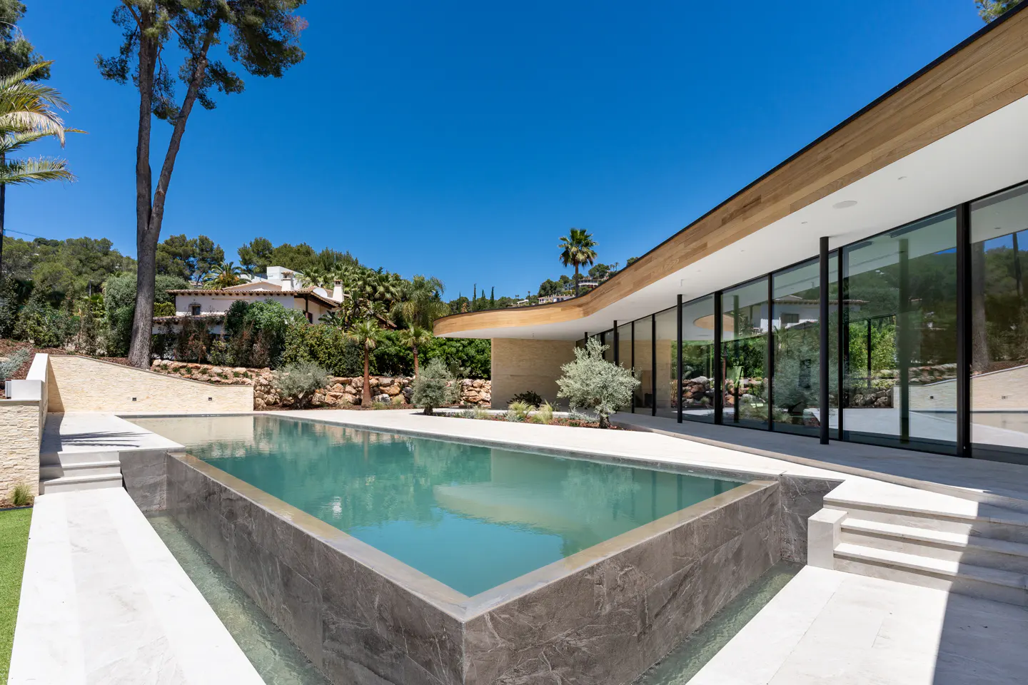 A modern home with a rectangular pool. The house has large glass windows and a light wood roof. Trees and blue sky are in the background.