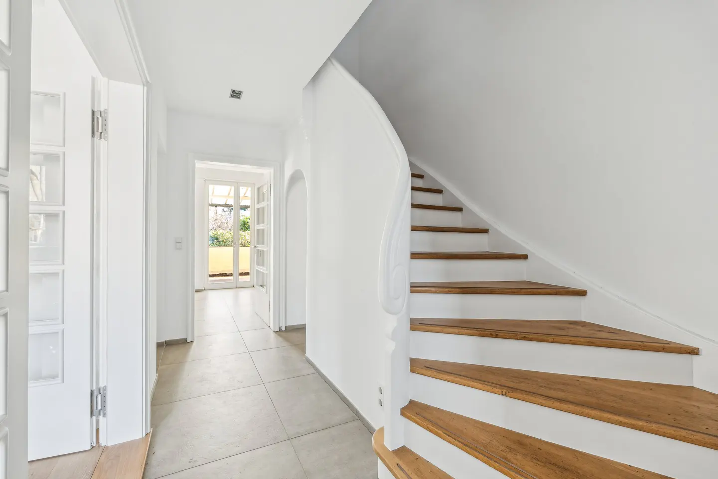 Bright hallway with white walls, tile floor, and wood-and-white stairs. A doorway leads to a sunlit patio.
