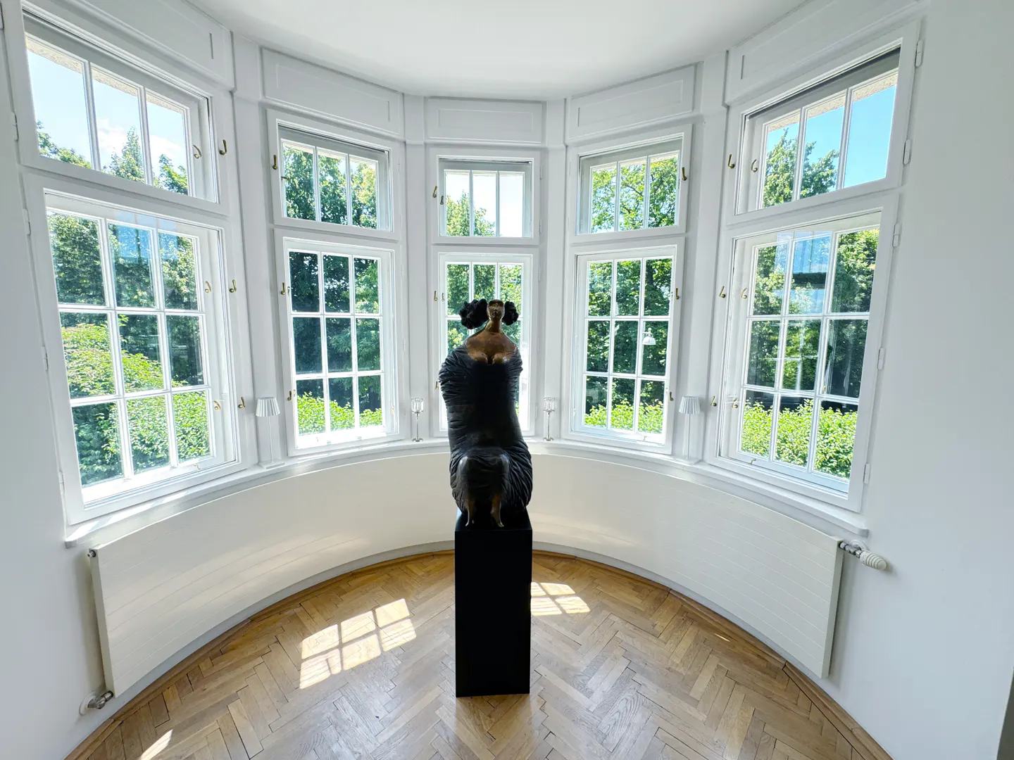 Bright room with parquet floors, white walls, and a curved bay window. A dark sculpture stands on a black pedestal in front of the windows.