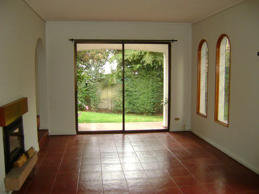 An empty living room with red tile floors, white walls, a fireplace, and a sliding glass door to a green backyard. Two arched windows are on the right.