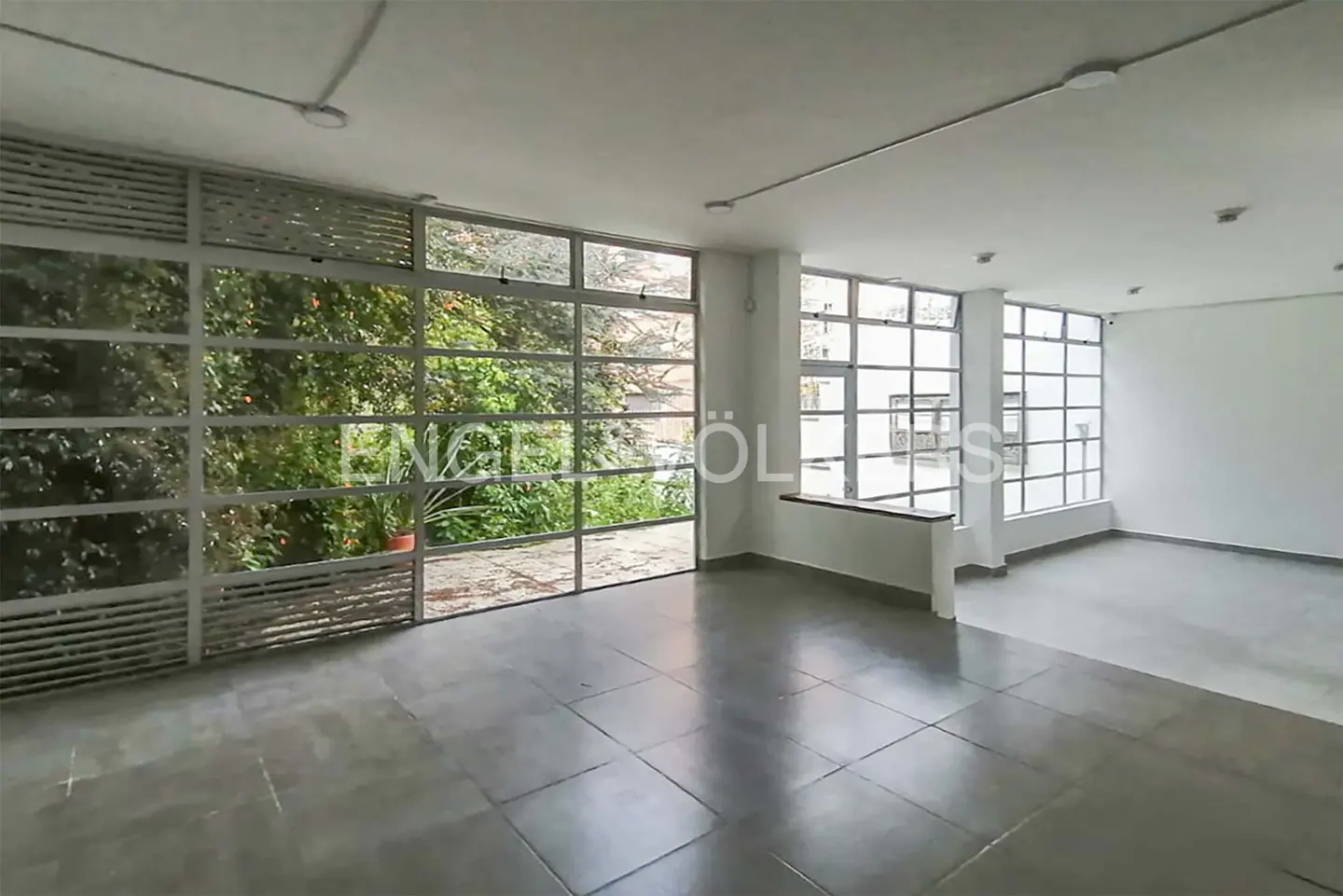 Empty room with gray tile floor and white walls. Large windows with a view of green foliage.