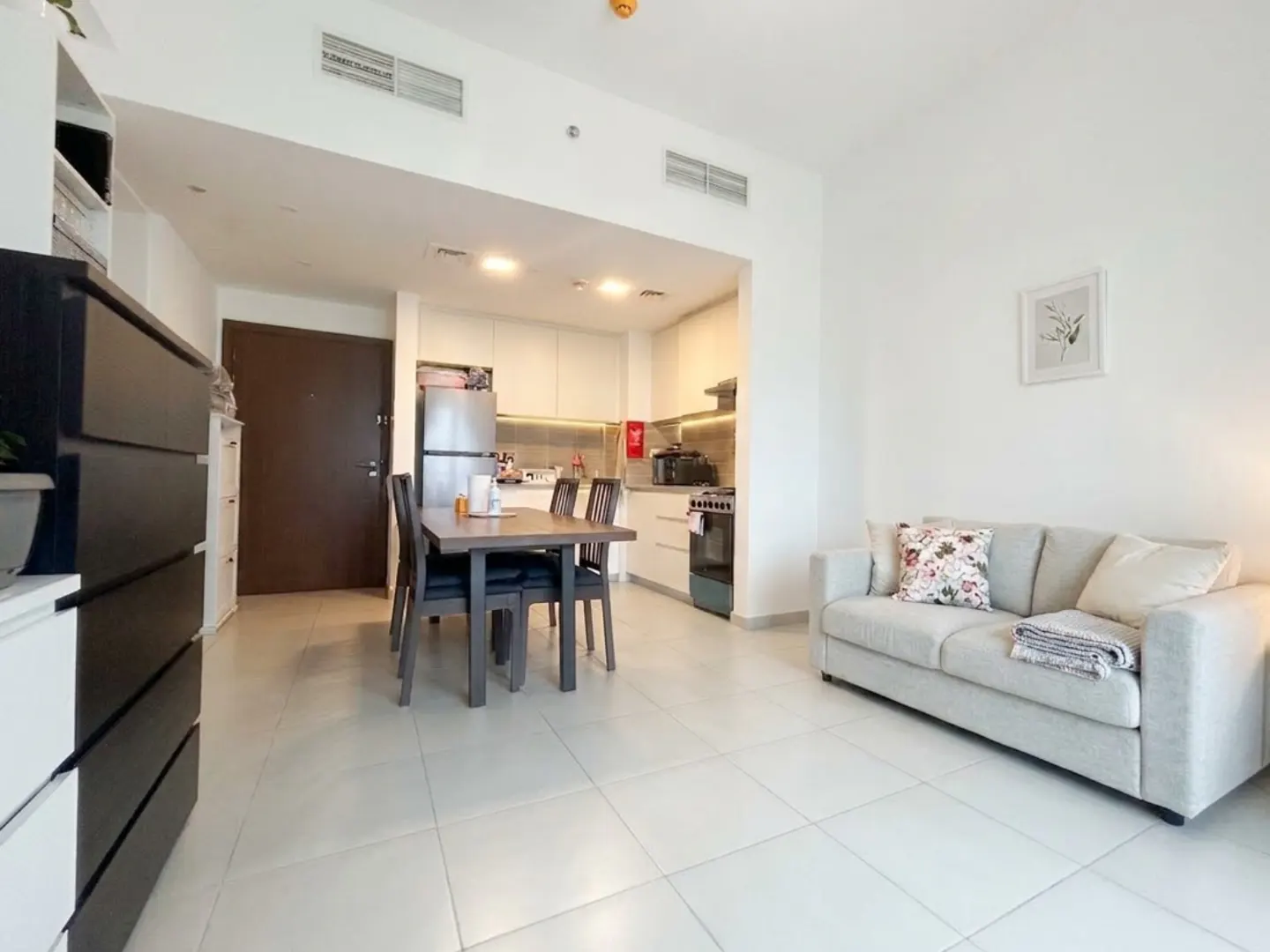 Bright apartment interior with a gray sofa, dark wood dining table, and white kitchen cabinets. Tiled floor and white walls create a clean, modern look.