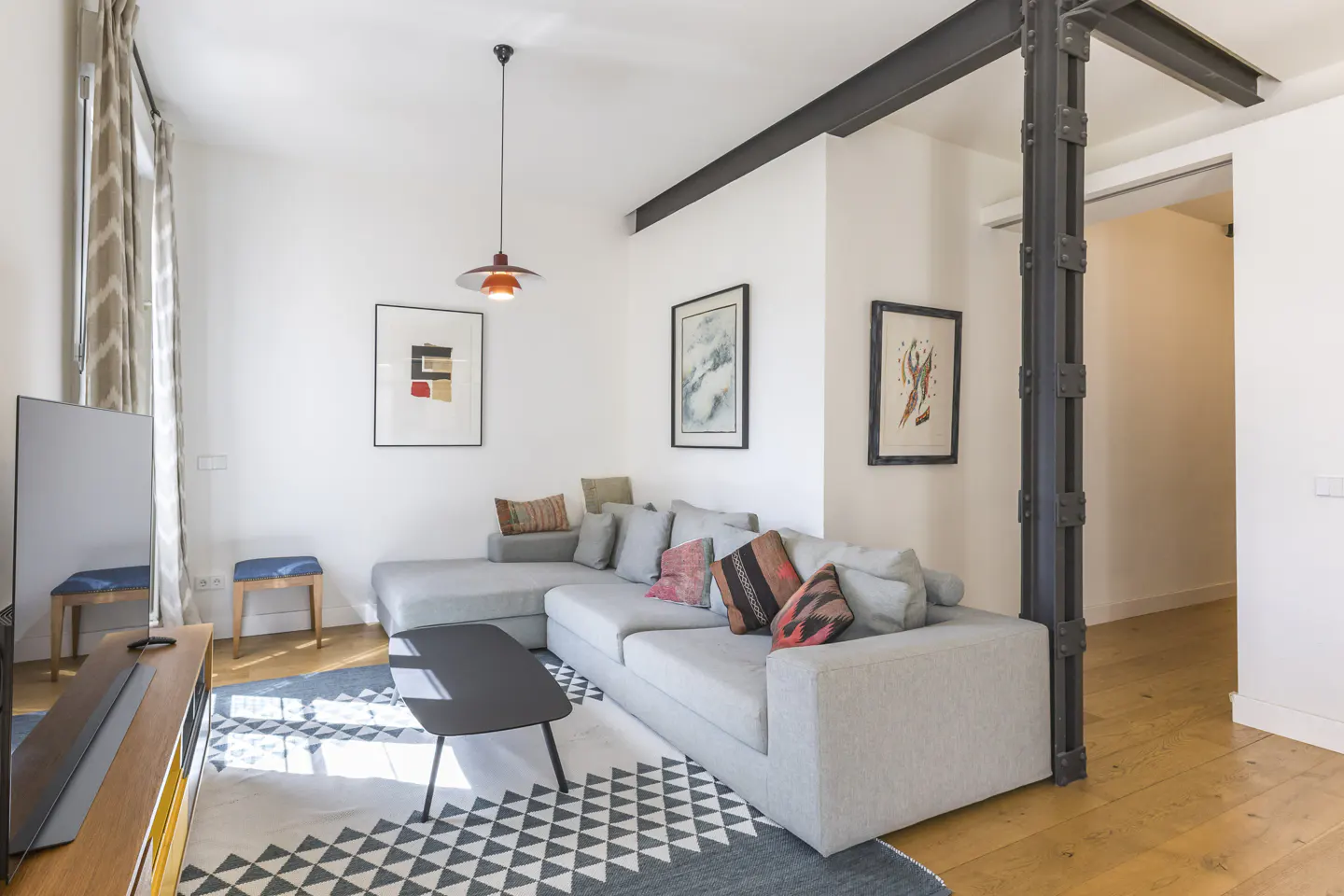 Bright living room with a gray sectional sofa, black coffee table, and geometric rug. Artwork adorns the white walls, and a black steel beam adds architectural detail.
