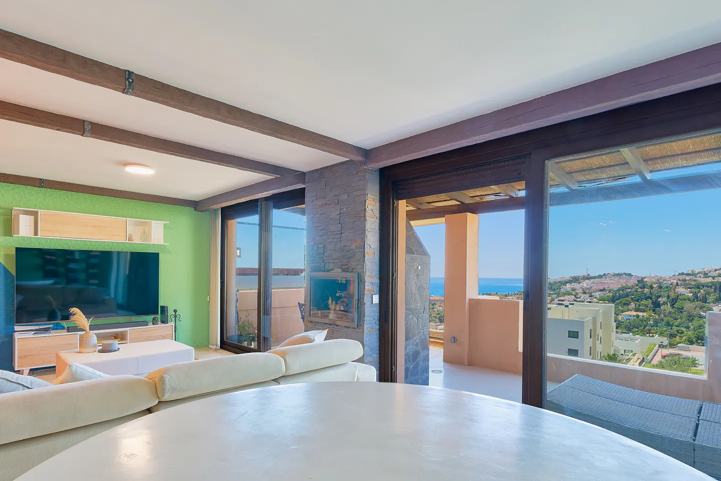Bright living room with green wall, white sofa, and large windows showing a coastal view. Beamed ceiling adds rustic charm.