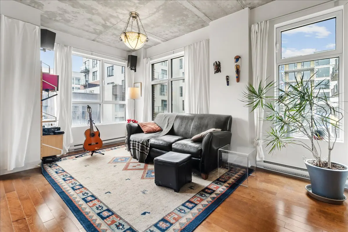Living room with hardwood floors, a gray leather sofa, and a patterned rug. Large windows offer city views. A guitar stands near a shelf.