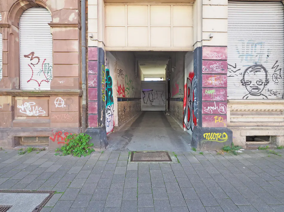 A narrow alleyway between two buildings, covered in graffiti, leads to a closed shutter in the distance.