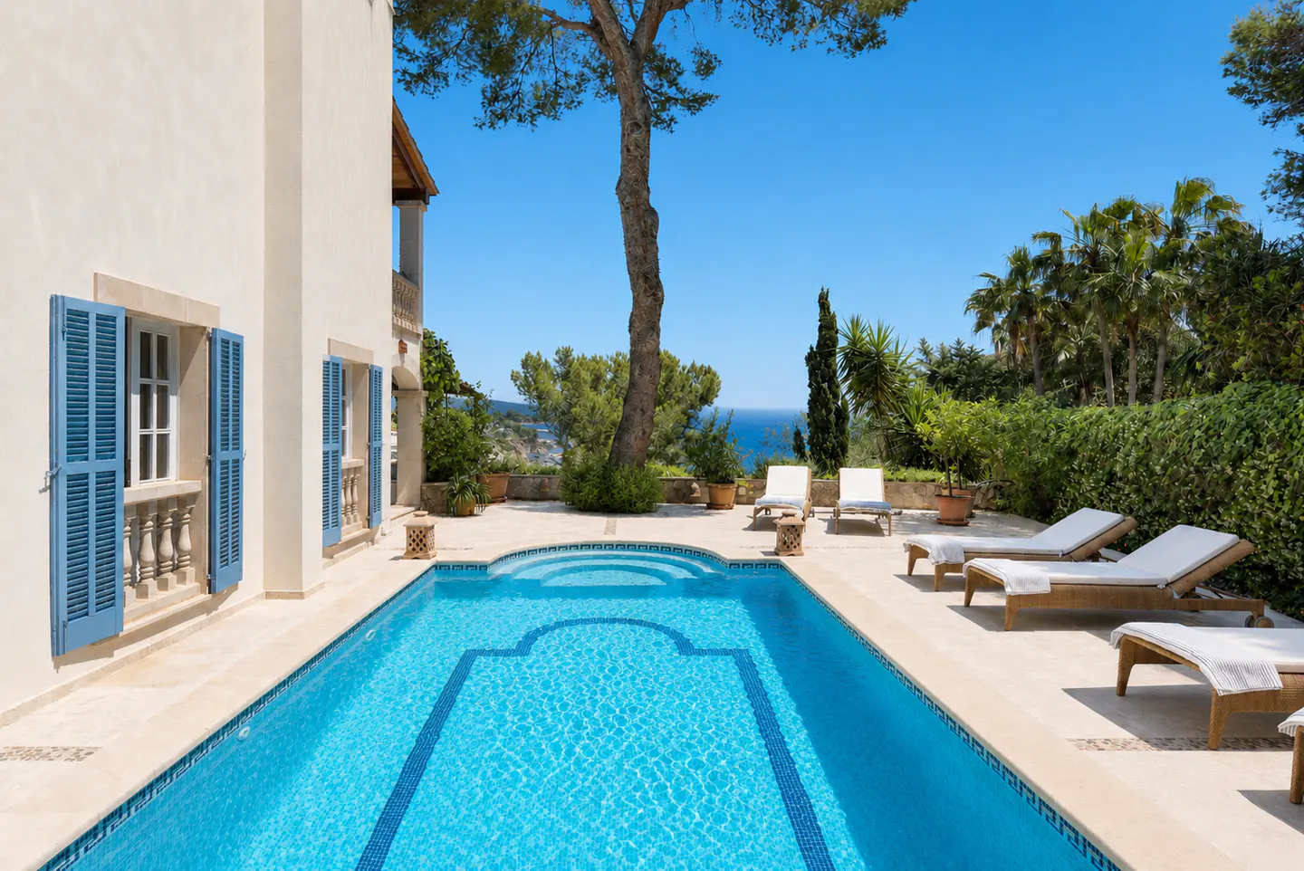 A swimming pool with blue water next to a cream-colored villa with blue shutters, lounge chairs, and a sea view.