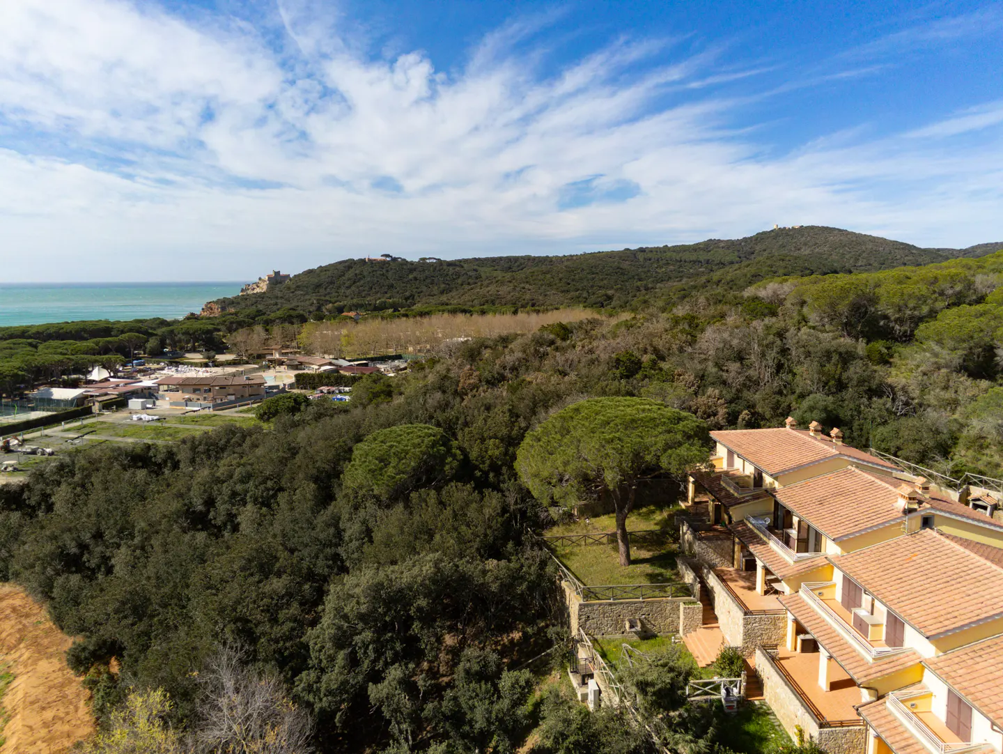 Scenic view of a yellow building with a red tile roof, surrounded by green trees, with the ocean in the background.