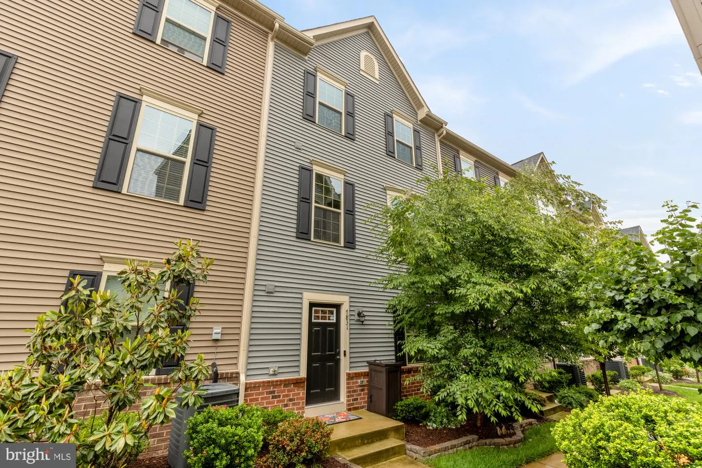 Exterior view of a two-story townhouse with gray siding, black shutters, and a black front door. Green bushes and trees surround the property.