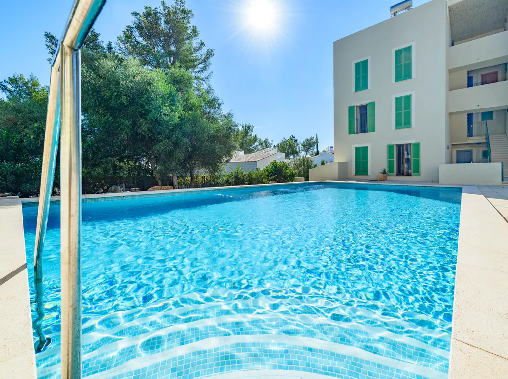 Outdoor pool with turquoise water, steps, and a metal handrail. A white building with green shutters is in the background.