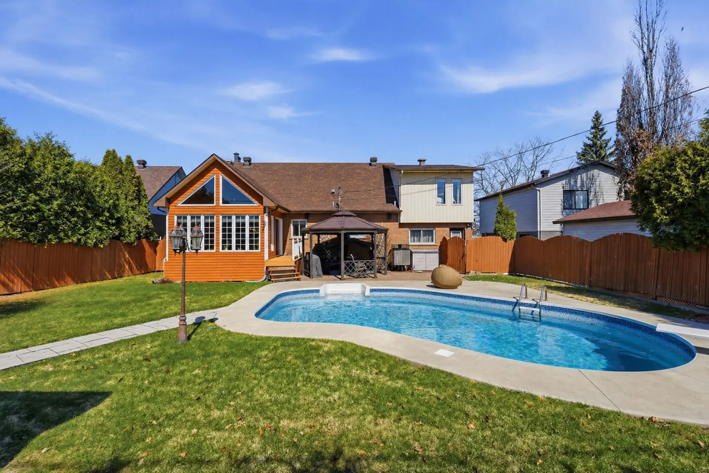 Backyard view of a house with a blue swimming pool, a gazebo, and a green lawn on a sunny day.