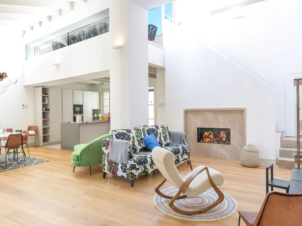 Bright, modern living room with hardwood floors, a colorful sofa, green chaise lounge, and a fireplace with a fire burning.