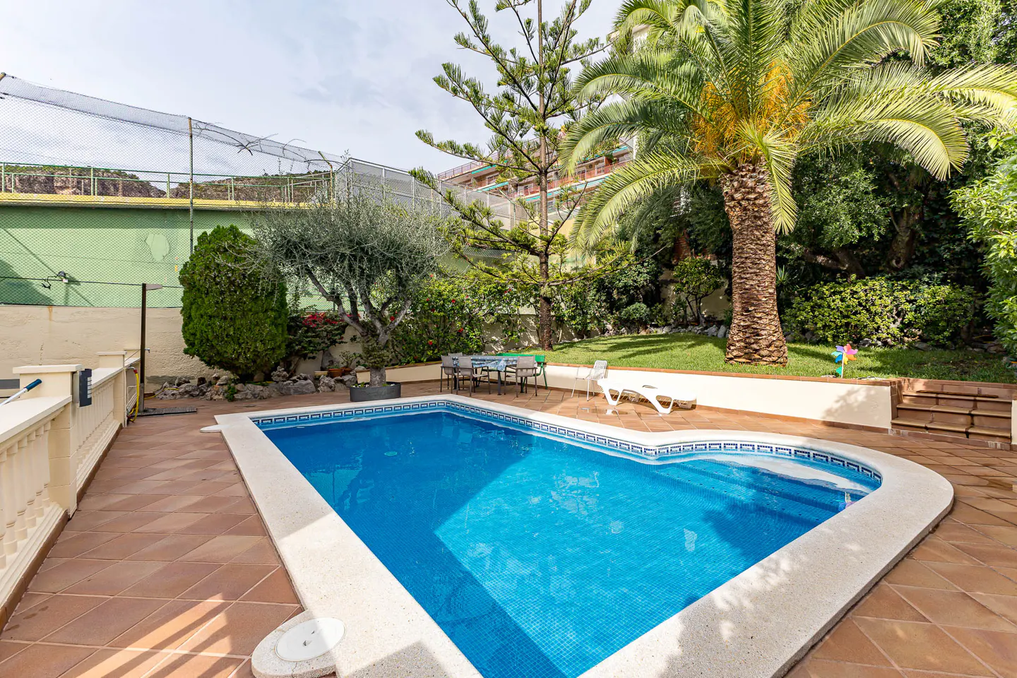 A backyard with a blue tiled pool, a table with chairs, a lounge chair, and lush green trees.