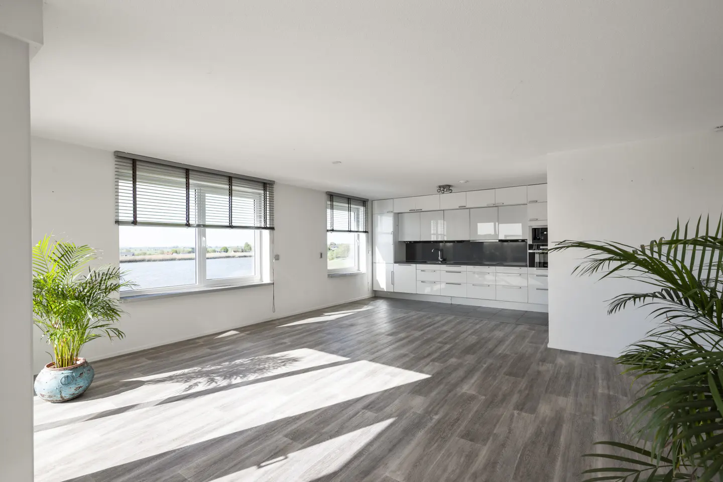Bright, open-concept apartment with gray wood floors, white walls, and a modern white kitchen. Windows offer water views. Potted plants add greenery.