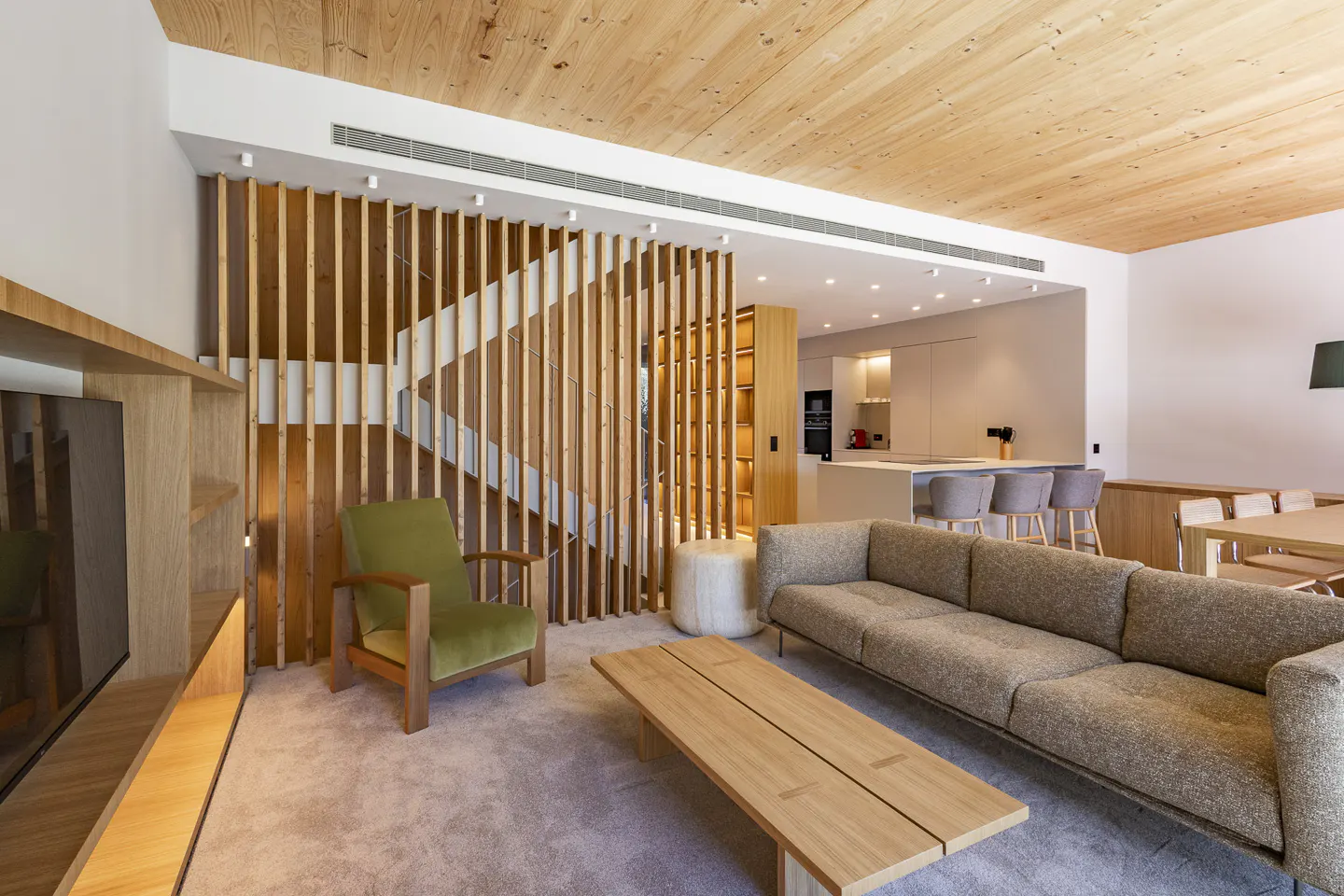 Modern living room with wood ceiling, gray sofa, and green chair. A wooden slat wall partially obscures the staircase. Kitchen in background.