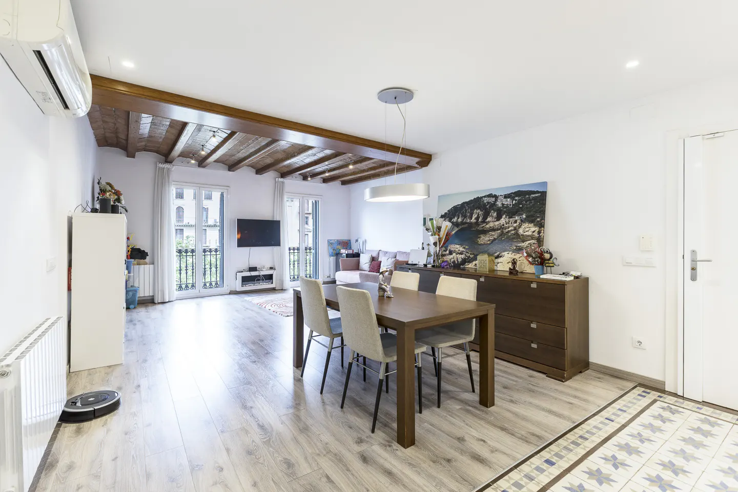 Bright, open living and dining area with wood floors, exposed beams, and balcony. Table with six chairs in the foreground.