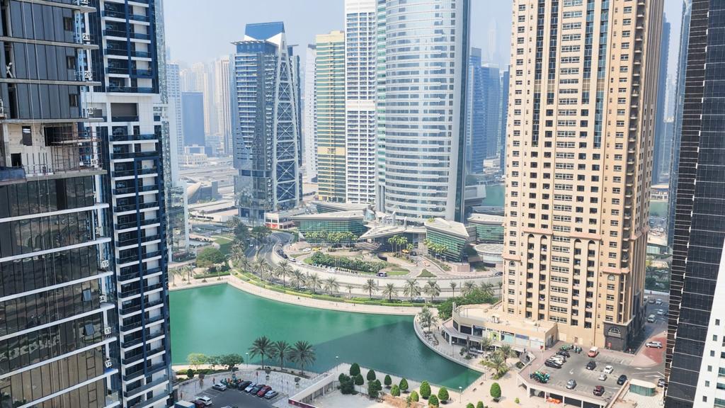 High-angle view of a modern city skyline with tall glass skyscrapers surrounding a central turquoise lake and palm trees. [AI-generated text]
