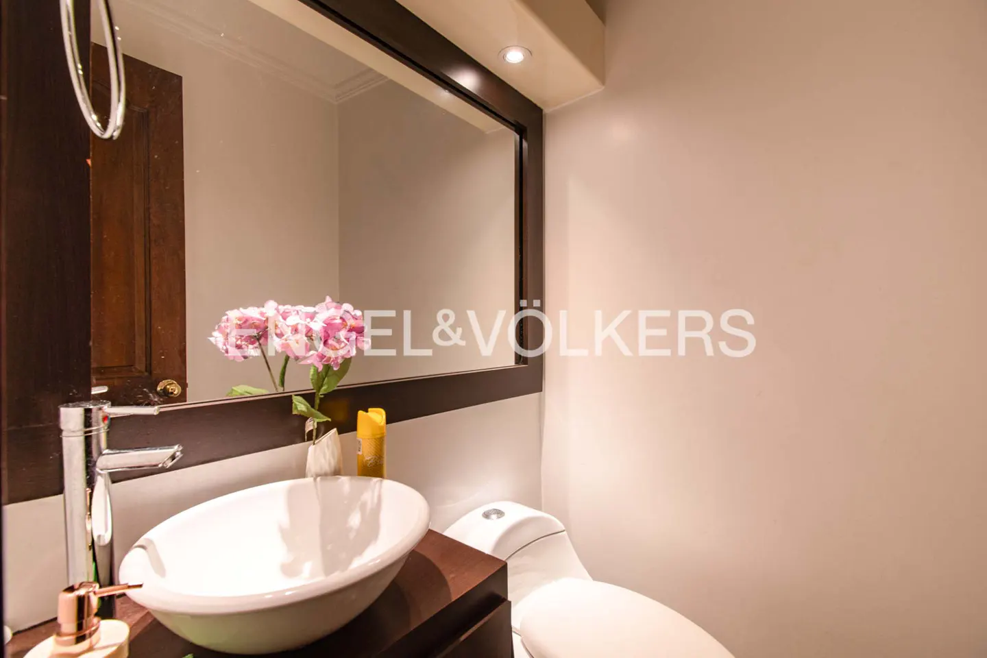 Bathroom with white sink, chrome faucet, and dark wood vanity. A mirror reflects pink flowers and a brown door. A white toilet is visible.