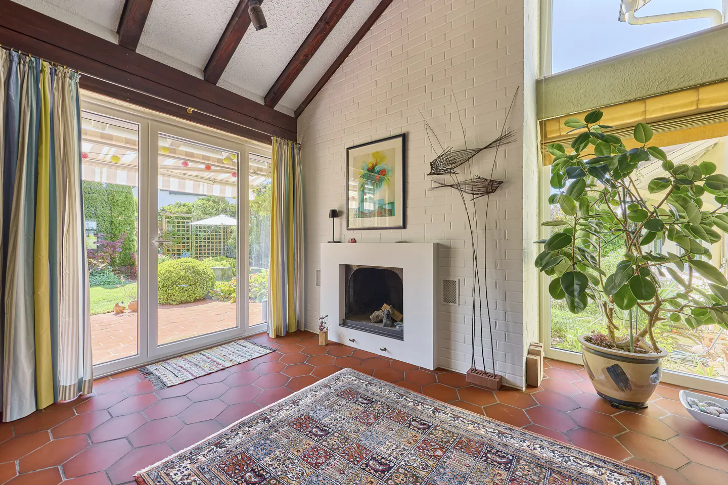 A sunroom with red tile floors, a white brick wall, and a fireplace. A patterned rug covers the floor. Curtains frame a glass door to a garden.