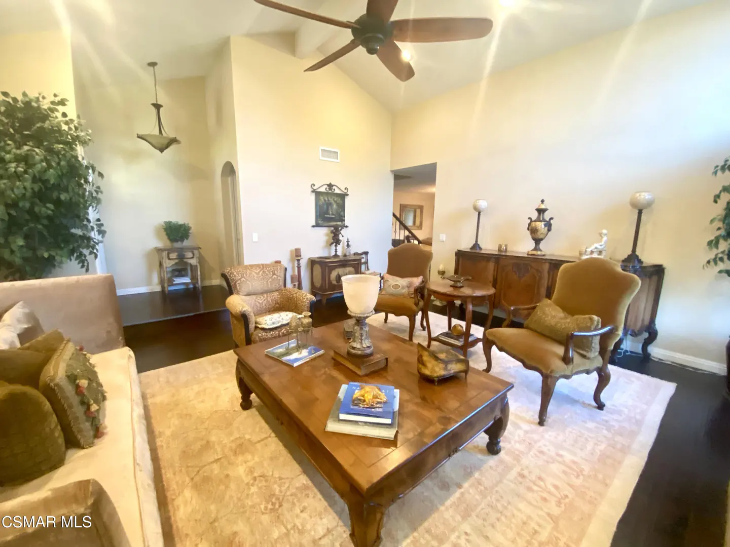A living room with tan walls, dark wood floors, and a ceiling fan. A large wooden coffee table sits on a light-colored rug, surrounded by chairs and cabinets.