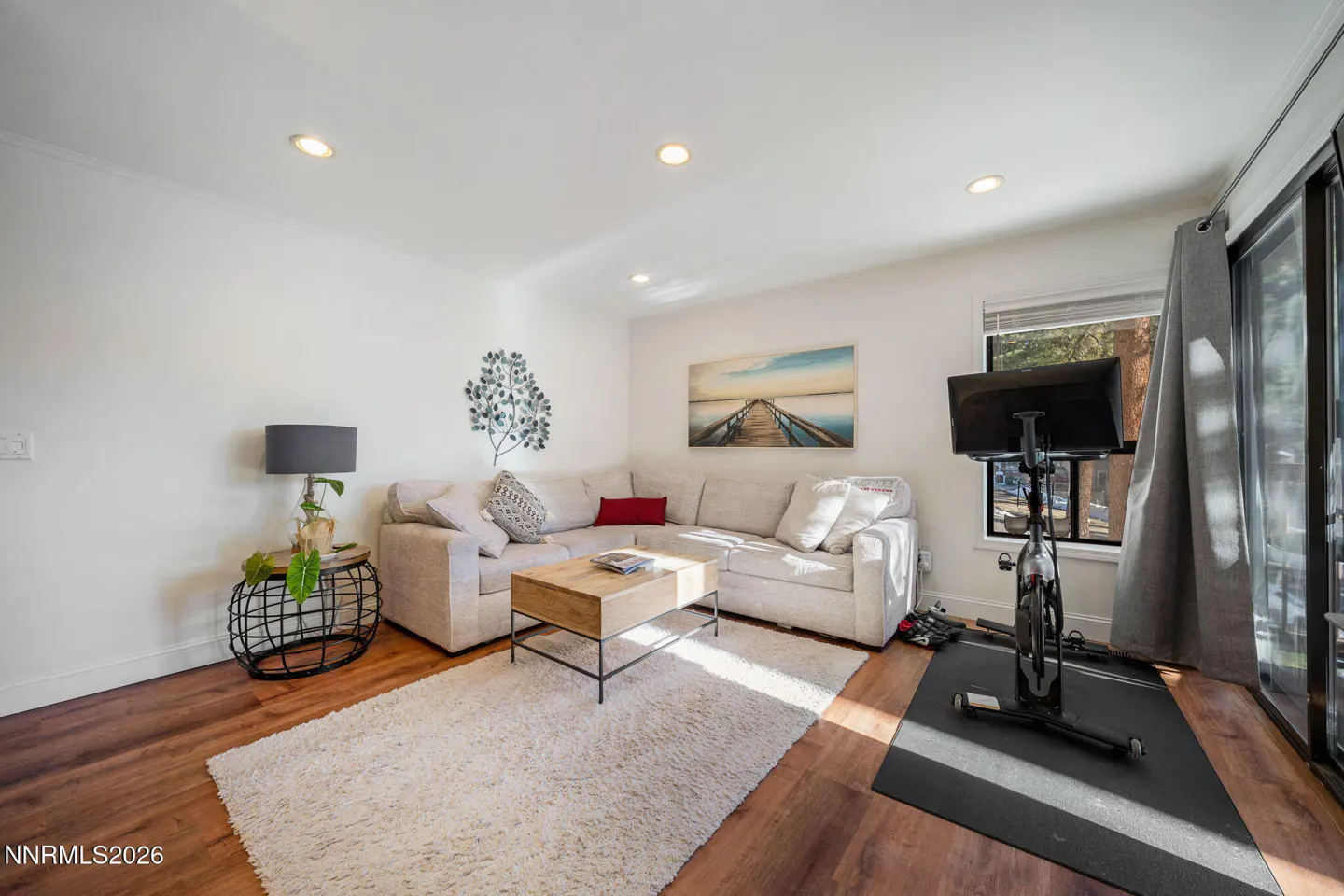 Living room with white walls, wood floors, and a large sectional sofa. A stationary bike sits near sliding glass doors. Artwork adorns the walls.