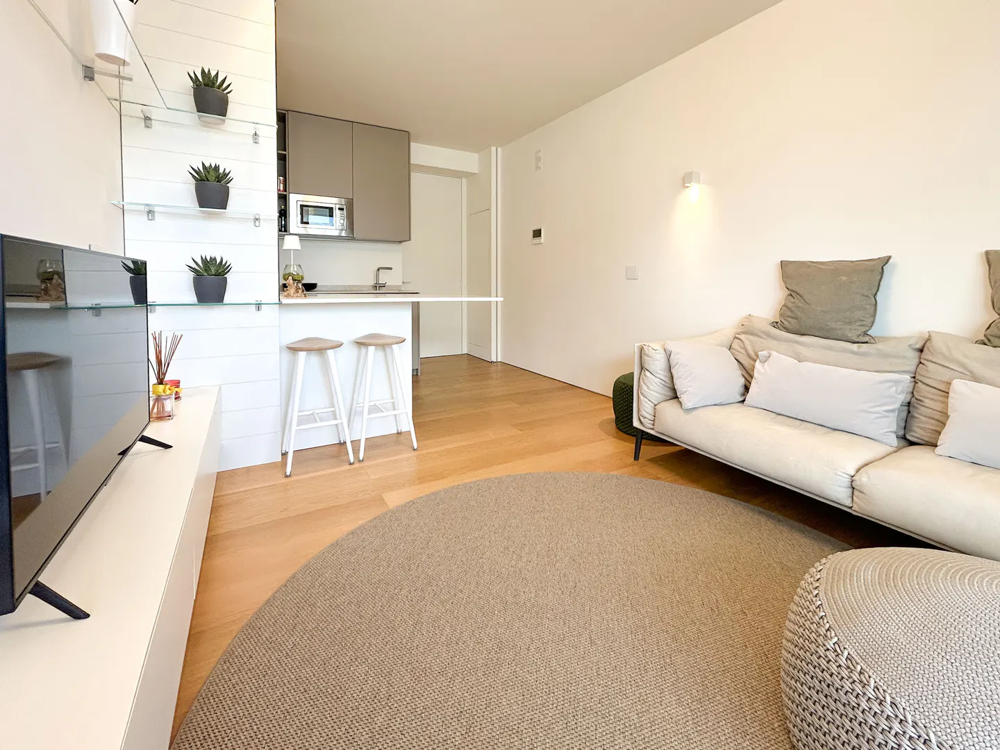 Bright, modern apartment with a beige round rug, white sofa, and kitchen with bar stools. A TV is on a white console.