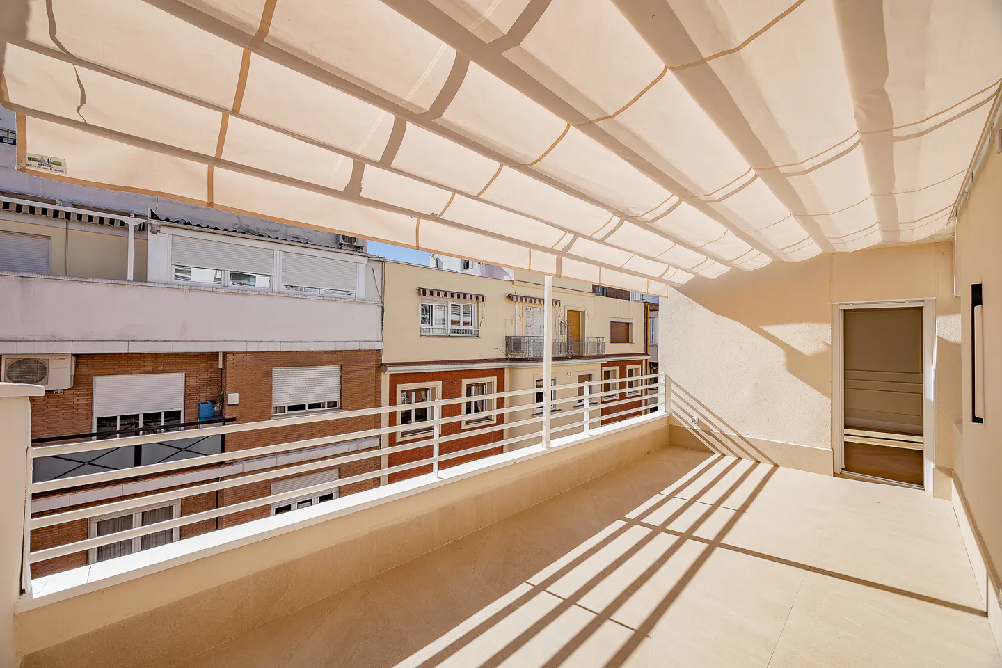A sunlit balcony with a retractable awning, white railings, and a view of neighboring buildings.
