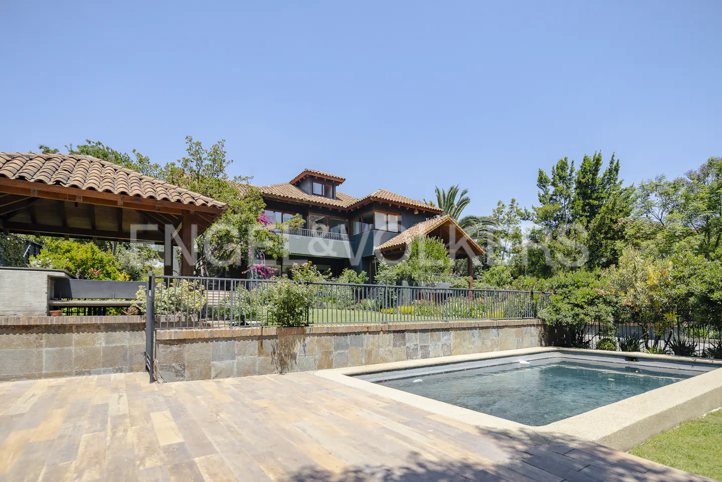 Backyard view of a two-story house with a pool, wooden deck, and outdoor kitchen under a clear blue sky.
