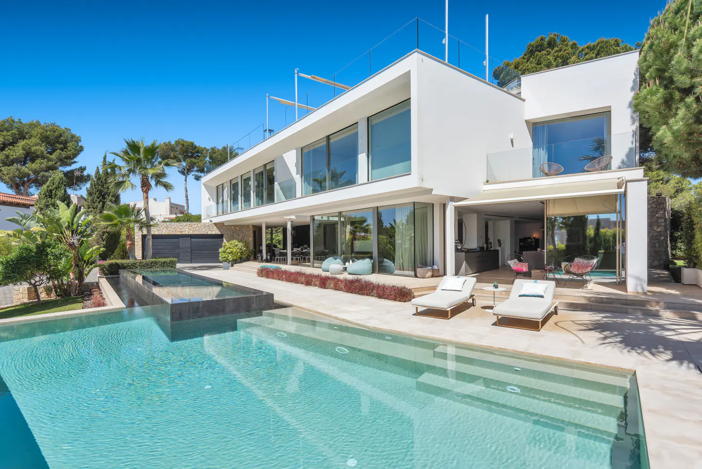 Modern white house with a pool. The pool has steps leading into the water. Lounge chairs are on the patio. Blue sky.