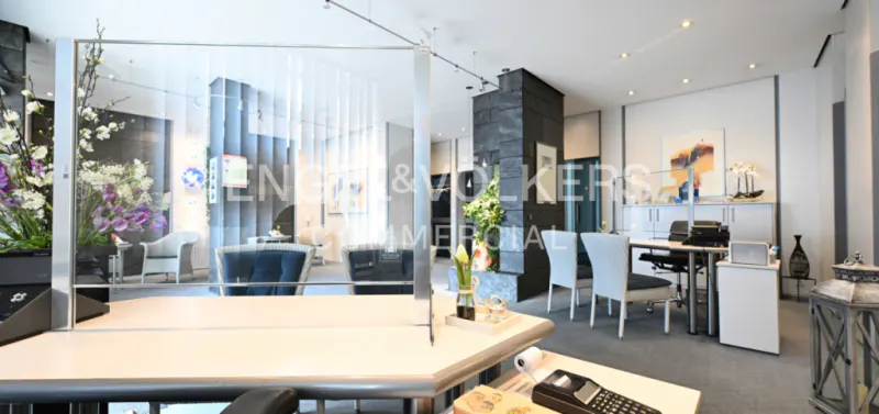 A modern real estate office with a reception desk, white chairs, and a lounge area with blue seating. Flowers add a touch of elegance.