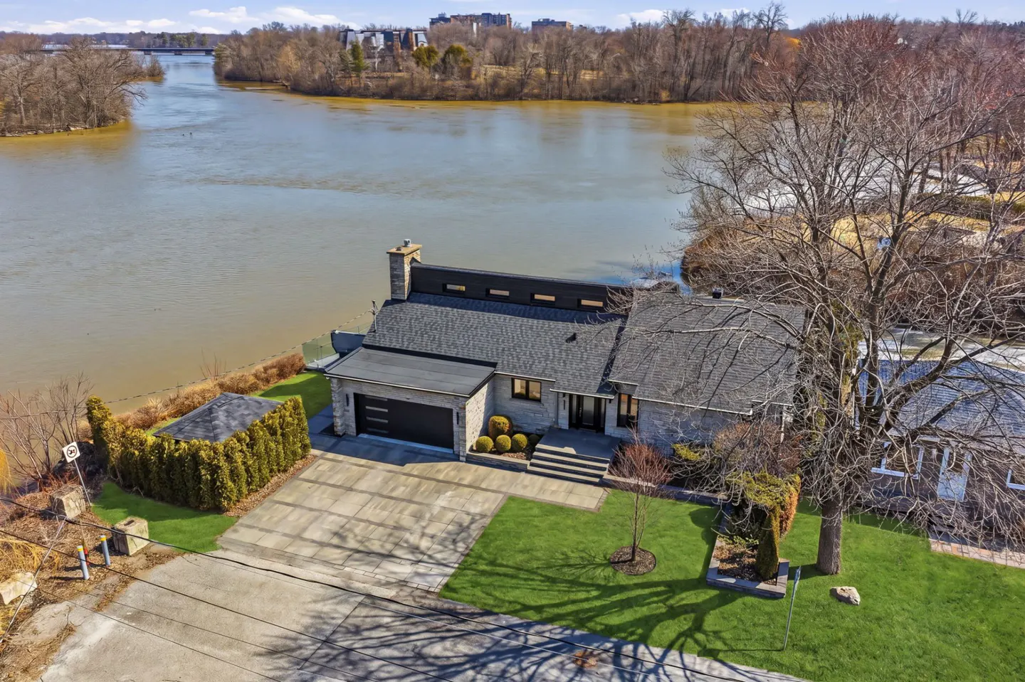 Aerial view of a stone house with a gray roof, a black garage door, and a river in the background.