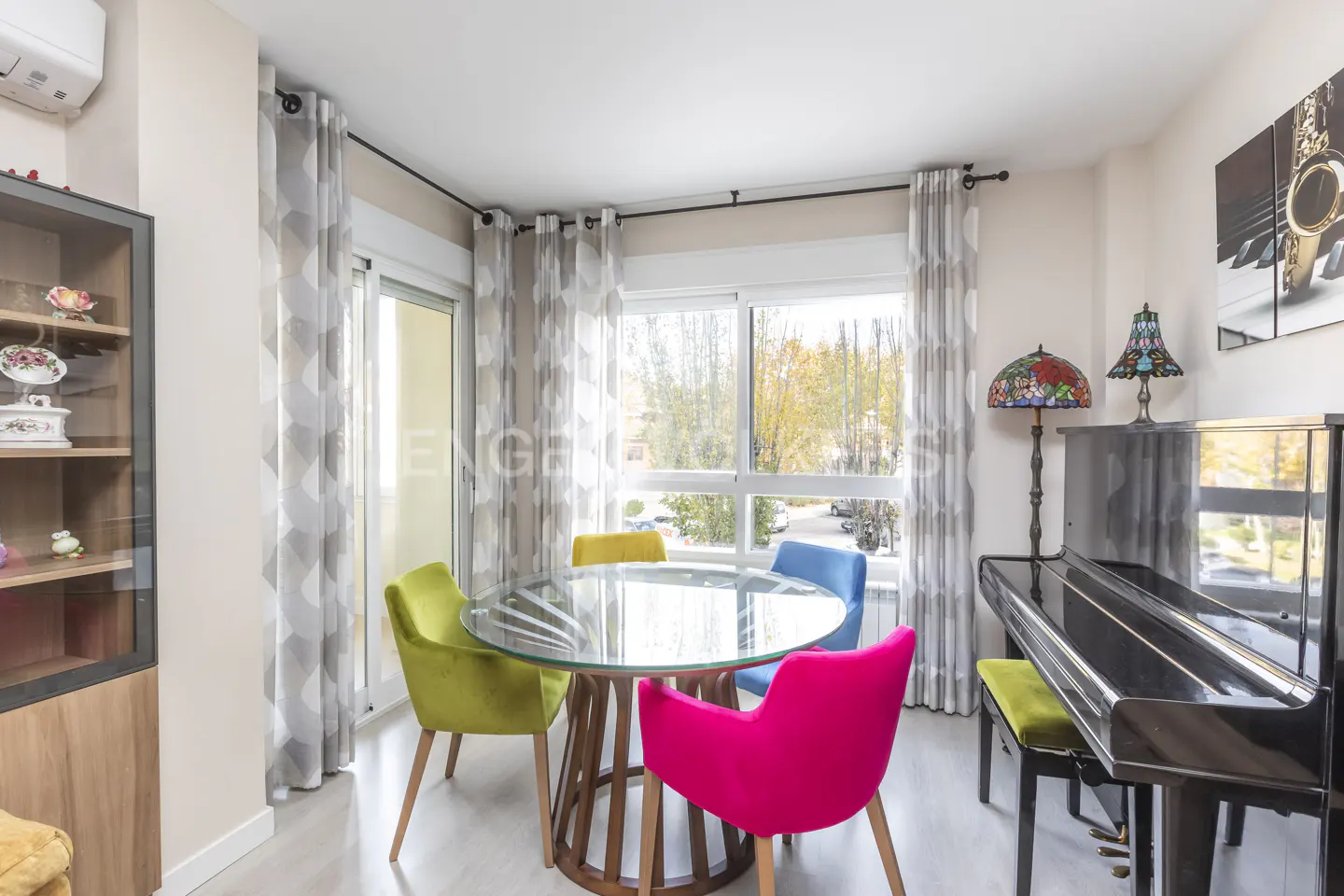 Bright dining area with a glass table, colorful chairs, and a black piano near a window with patterned curtains.