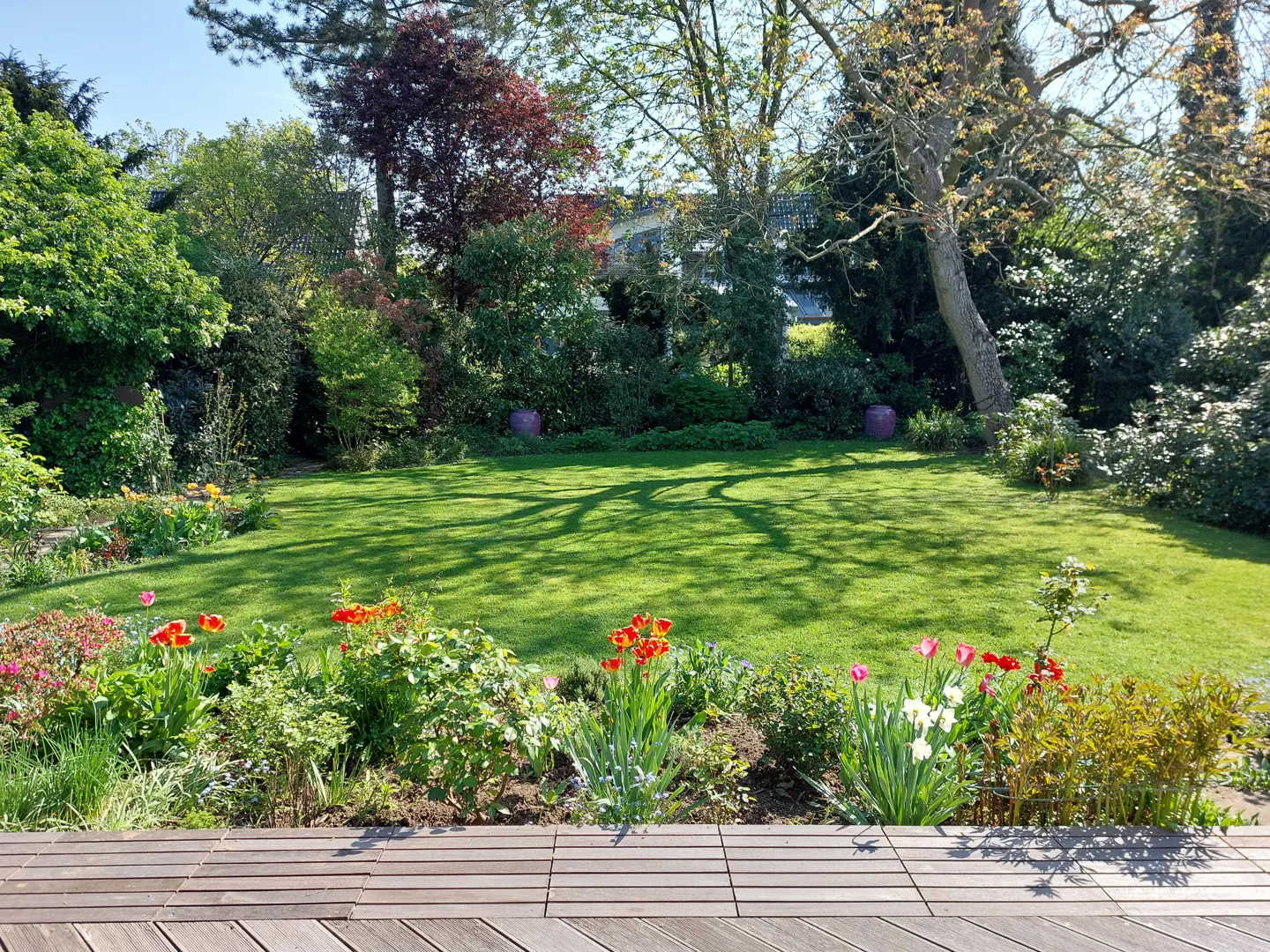 Lush green lawn with flower bed, viewed from a wooden deck. Trees and shrubs surround the yard.