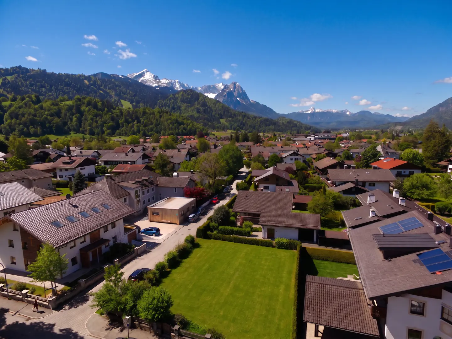 Aerial view of a European town with brown-roofed houses, green trees, and snow-capped mountains under a blue sky.