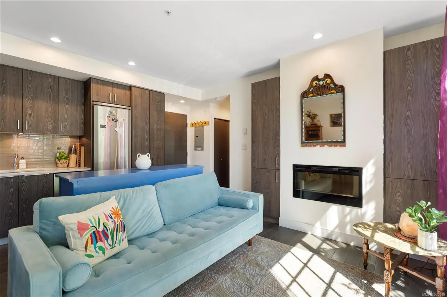 Living room with a light blue sofa, a blue kitchen island, and dark wood cabinets. A fireplace and mirror are on the right.