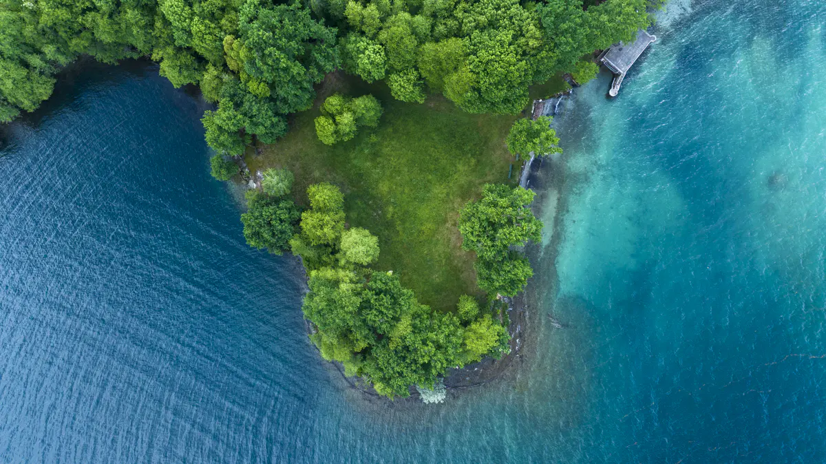 Aerial view of a green island surrounded by turquoise and dark blue water. A small dock is visible on one side.