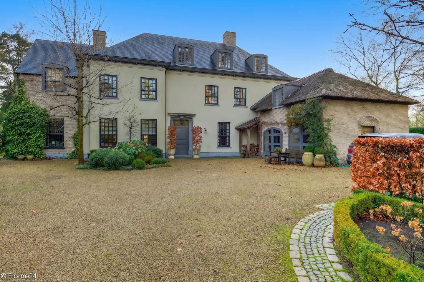 Two-story house with a gray roof, cream-colored walls, and black-framed windows, surrounded by a gravel driveway and manicured greenery.