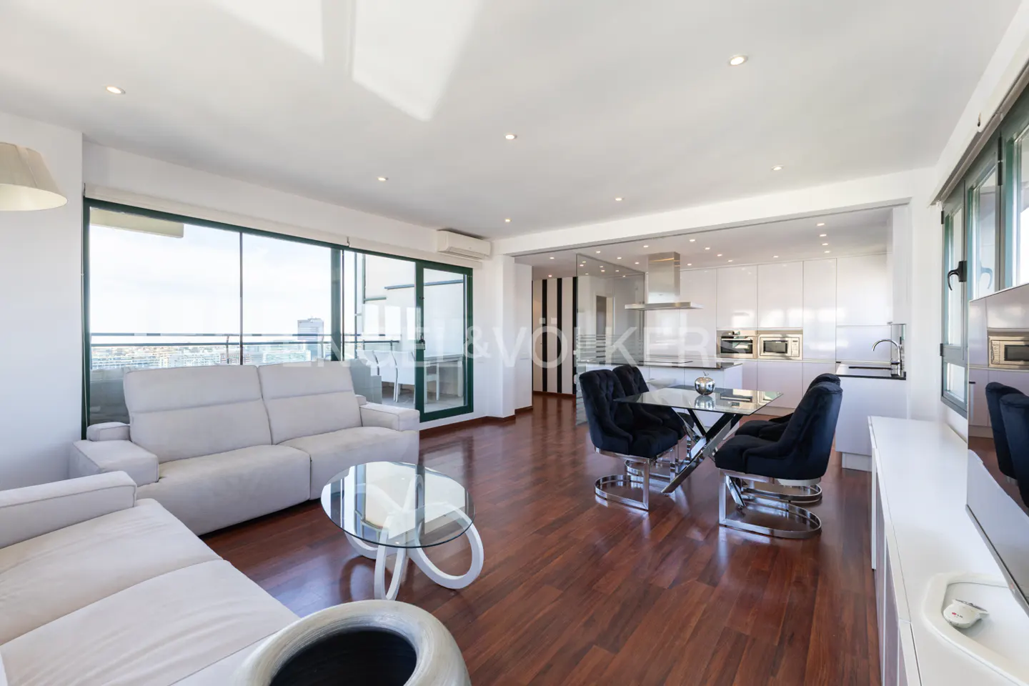 Bright, modern apartment with white sofas, glass tables, and black dining chairs. Open kitchen with white cabinets and stainless steel appliances. Balcony with city view.