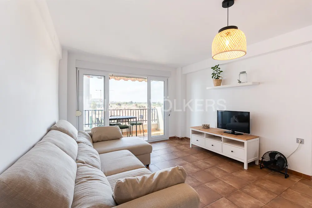 Bright living room with a beige sectional sofa, TV, and balcony with outdoor seating. The floor is tiled in brown.