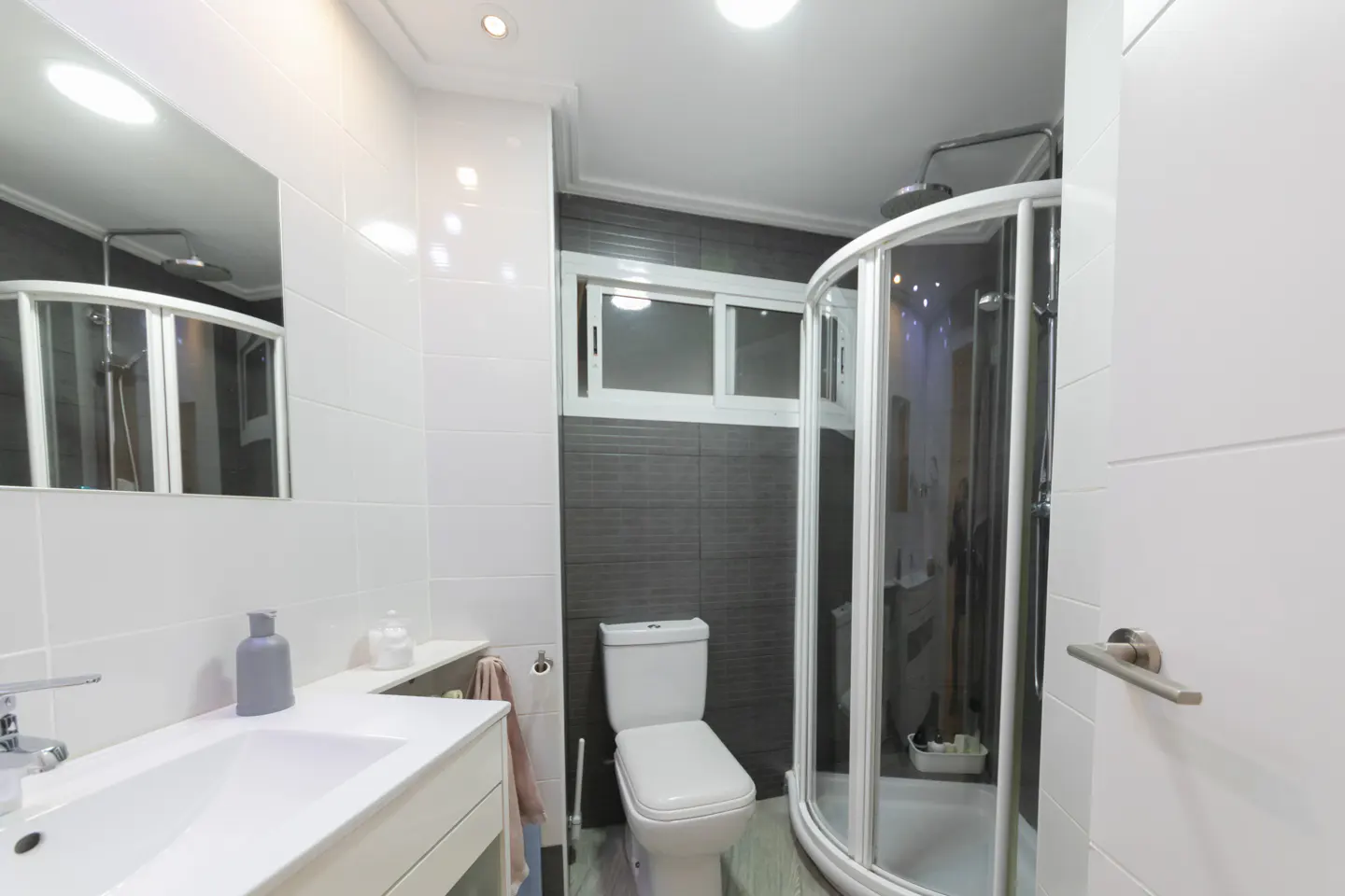 Bright bathroom with white sink, toilet, and curved glass shower. Gray tiled wall and white door visible.
