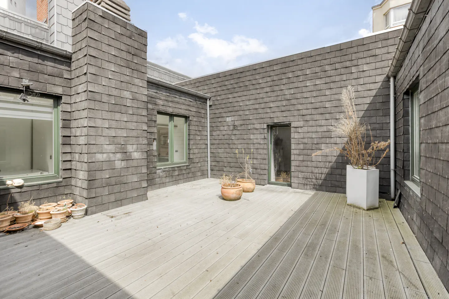 Exterior shot of a rooftop deck with gray wood planks and gray shingle siding. Potted plants are scattered around the deck.