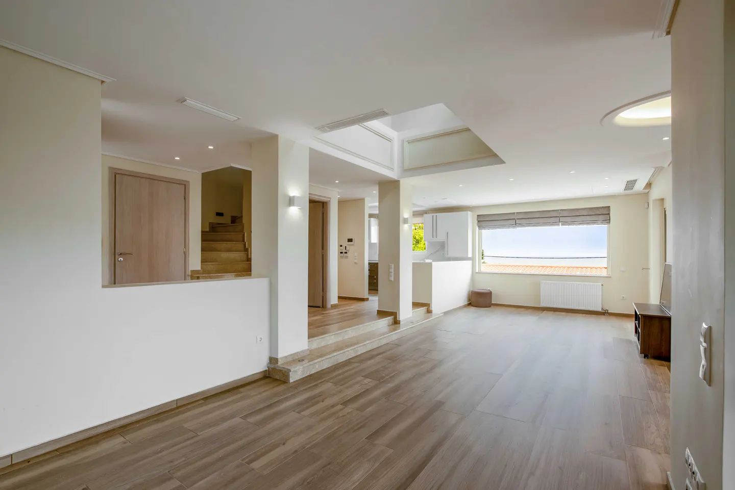 Spacious, modern living area with light wood floors, a sunken floor level, white walls, and a large window with a view.