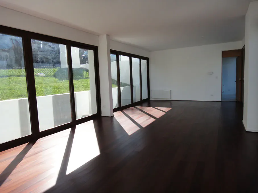 Bright, empty room with dark wood floors and white walls. Large windows show a green lawn outside. Sunlight streams in.