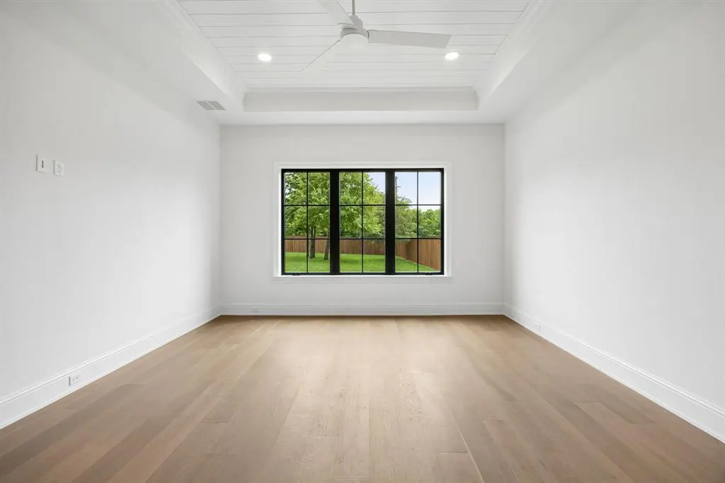 Bright, empty room with white walls, light wood floors, and a black-framed window showing a green lawn and trees. A white ceiling fan is centered above.