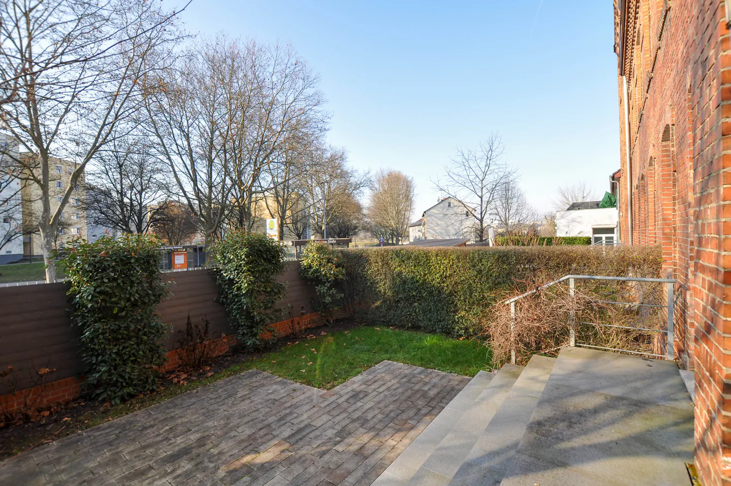Brick patio with steps leading to a small lawn, bordered by a hedge and a brown fence. Bare trees and buildings in the background.