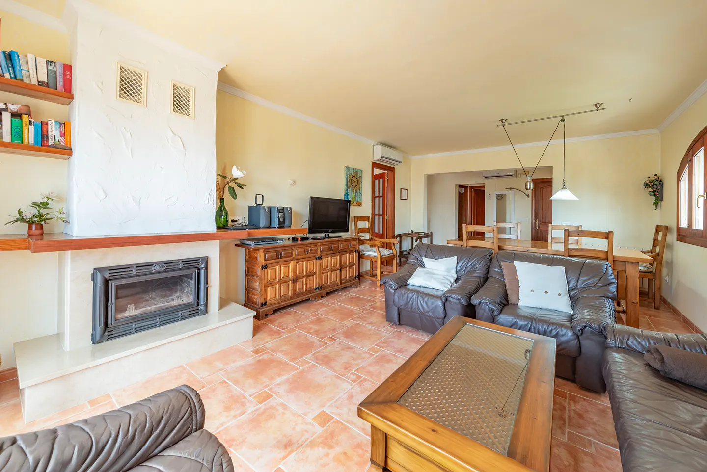 A living room with brown leather sofas, a wooden coffee table, and a fireplace. A dining table is visible in the background.