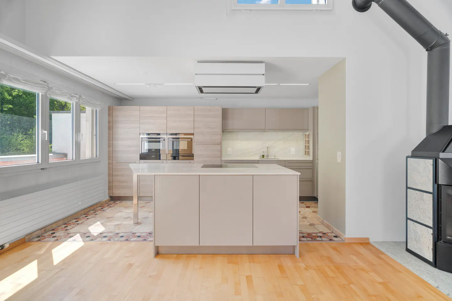 A bright, modern kitchen with light wood cabinets, a white island, and a black fireplace. Sunlight streams through windows.