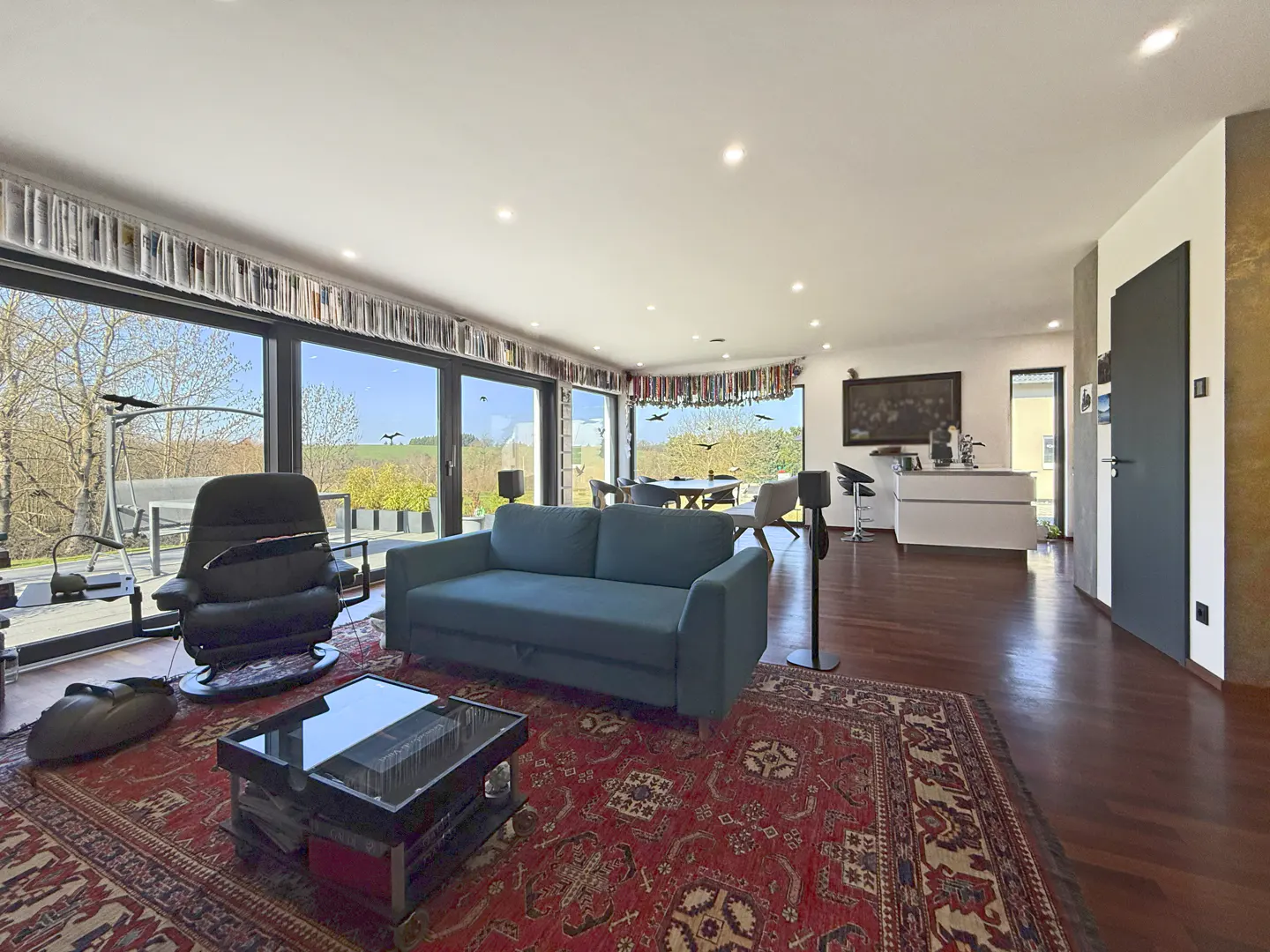 Bright living room with a blue sofa, black chair, and red rug. Large windows offer a view of the outdoors. Books line the top of the windows.