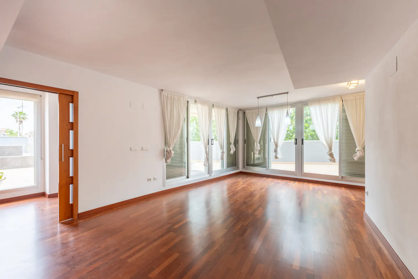 Bright, empty room with hardwood floors, white walls, and multiple glass doors with white curtains leading to a balcony.